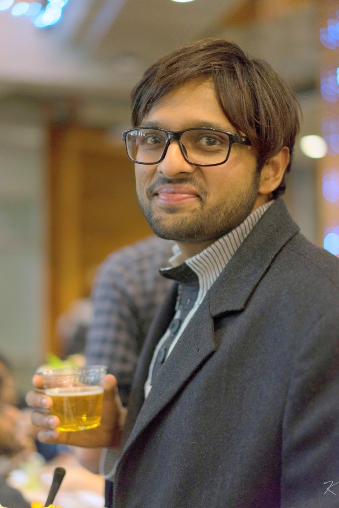A man with short dark hair and glasses holds a clear plastic cup containing a light‐colored beverage. He is looking toward the camera with a slight smile, wearing a dark coat and a collared shirt, with soft, out‐of‐focus lights in the background.